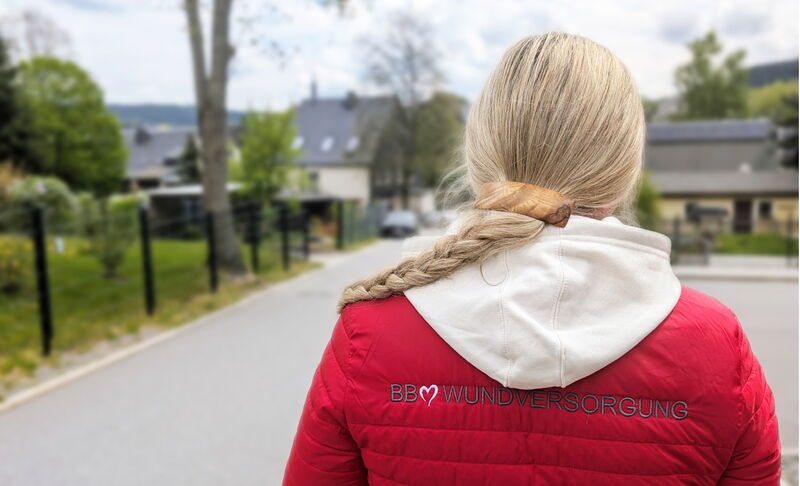 Eine Frau mit geflochtenem Zopf und roter Jacke mit dem Logo „BB Wundversorgung“ auf dem Rücken steht auf einer Strasse mit Blick auf ein Wohngebiet.