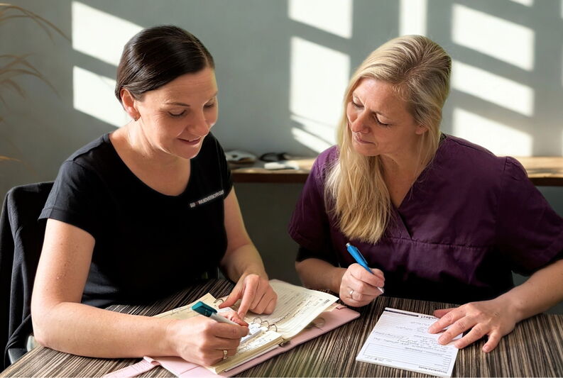 Zwei Frauen sitzen an einem Tisch und arbeiten gemeinsam an Formularen. Die Frau links trägt ein schwarzes Shirt mit der Aufschrift „BB Wundversorgung“ und schreibt in einen Ordner. Die Frau rechts trägt ein dunkelviolettes medizinisches Oberteil und hält
