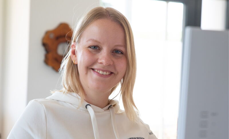 Eine junge Frau mit blondem Haar trägt eine weisse Kapuzenjacke und steht in einem Innenraum. Sie lächelt freundlich in die Kamera.