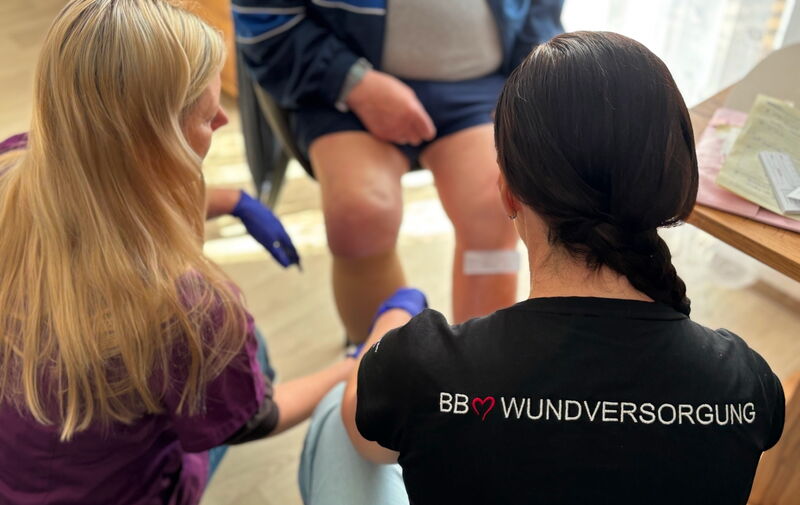 Zwei Pflegefachpersonen versorgen die Beine einer sitzenden Person. Beide tragen Handschuhe; eine trägt ein schwarzes T-Shirt mit der Aufschrift „BB WUNDVERSORGUNG“, die andere ein violettes Oberteil. An den Beinen der sitzenden Person sind weisse Verbänd