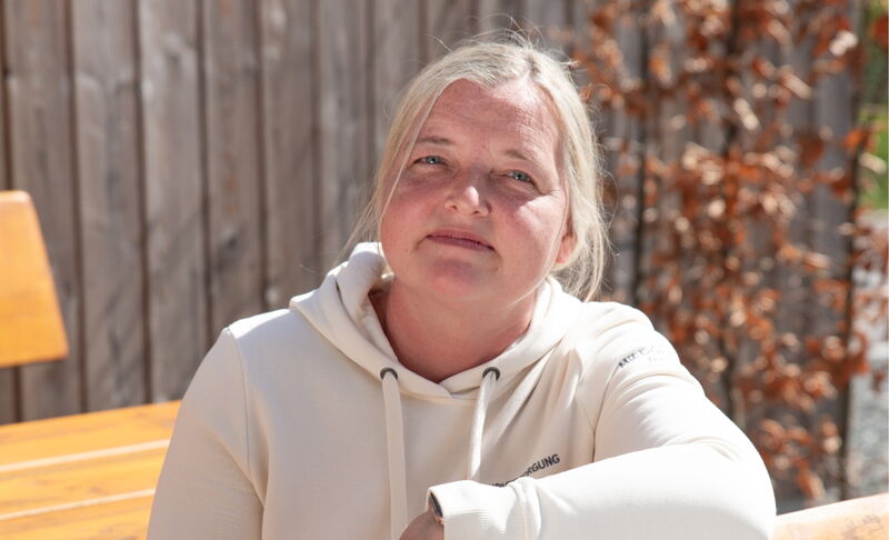 Eine Frau mit blonden Haaren in heller Kapuzenjacke sitzt an einem Holztisch vor einem Holzzaun und blickt ruhig in die Kamera. Im Hintergrund ist ein Strauch mit braunem Laub sichtbar.