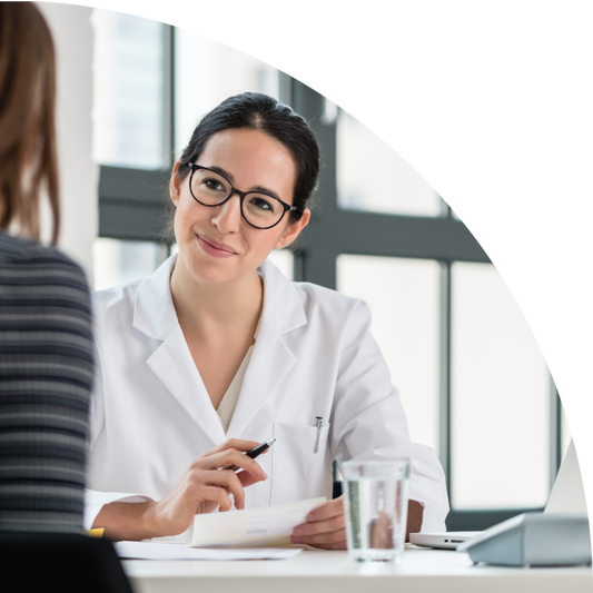 Eine Ärztin mit dunklen Haaren, Brille und weissem Kittel sitzt an einem Schreibtisch und führt ein Gespräch mit einer Patientin. Die Ärztin hält ein Formular und einen Stift in der Hand und lächelt freundlich. Im Vordergrund sind ein Glas Wasser und ein