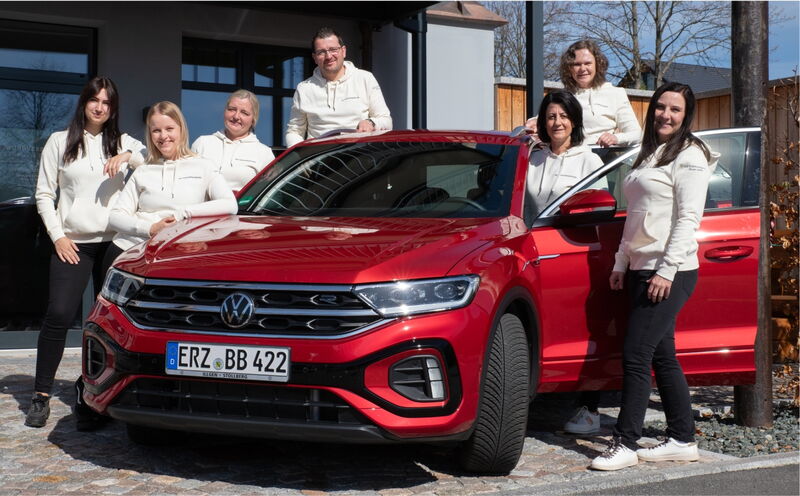 Sieben Personen stehen in weissen Kapuzenpullovern um ein rotes VW-SUV mit deutschem Kennzeichen „ERZ BB 422“. Das Auto steht auf einem gepflasterten Platz vor einem modernen Gebäude mit Glastür. Die Gruppe wirkt fröhlich und posiert gemeinsam bei Sonnens