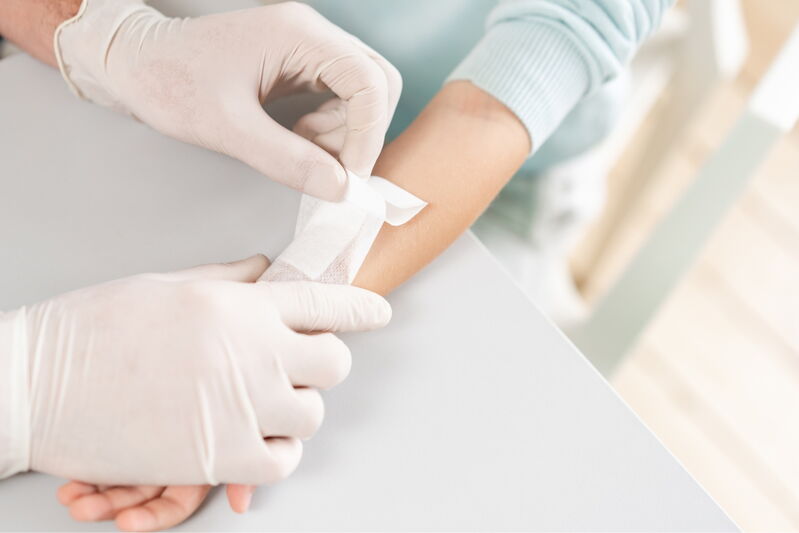 Eine Person mit weissen medizinischen Handschuhen legt einem Kind eine weisse Mullbinde am Unterarm an. Der Arm liegt auf einer weissen Tischplatte, im Hintergrund ist ein heller Raum erkennbar.