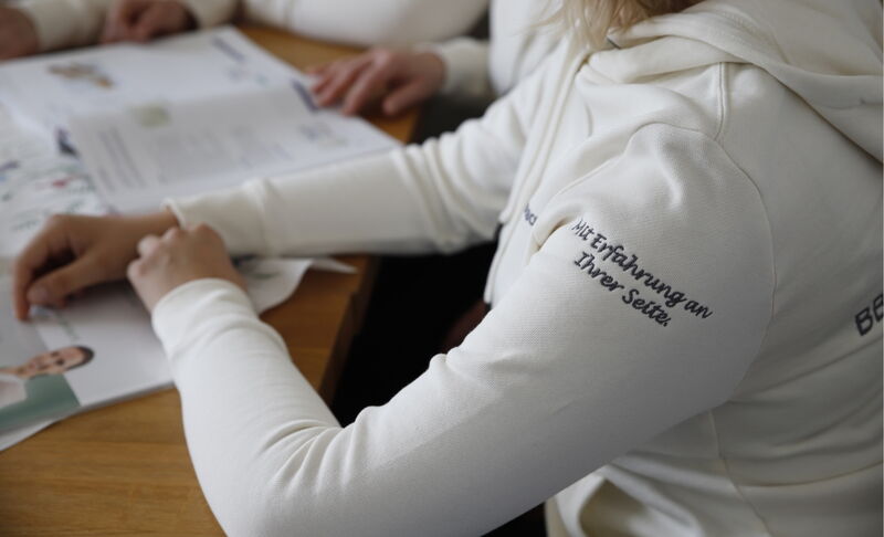 Mehrere Personen in weisser Kleidung mit dem Schriftzug „Mit Erfahrung an Ihrer Seite“ am Ärmel sitzen an einem Tisch und arbeiten mit gedruckten Unterlagen.