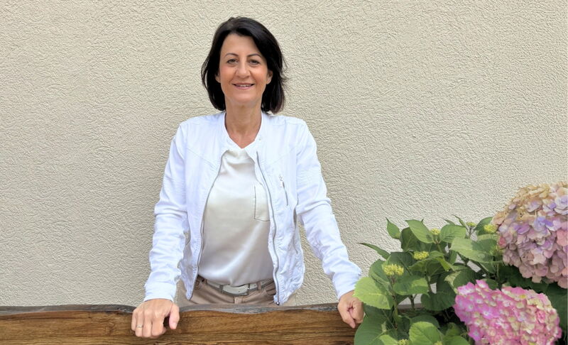 Eine Frau in weisser Jacke steht lächelnd hinter einem Holzbalken vor einer beigen Wand mit blühenden Hortensien im Vordergrund.