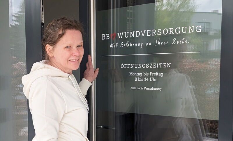 Eine Frau in heller Kapuzenjacke öffnet die Tür einer Praxis mit der Aufschrift "BB Wundversorgung – Mit Erfahrung an Ihrer Seite". Darunter stehen die Öffnungszeiten.